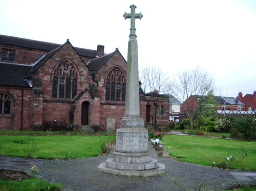 War memorial in Ashton-in-Makerfield, featuring a tall stone cross surrounded by greenery and church architecture in the background, symbolising local heritage and remembrance.