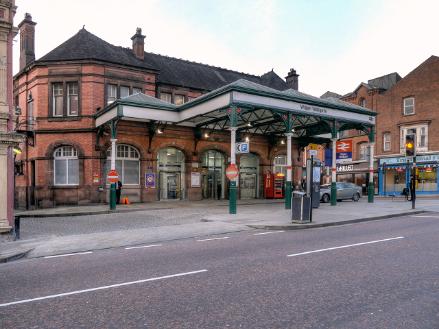 Wigan Wallgate train station entrance with distinctive red brick architecture, nearby parking signs, and local shops visible in the background, representing JJK Gas Services' commitment to serving the Wigan community.